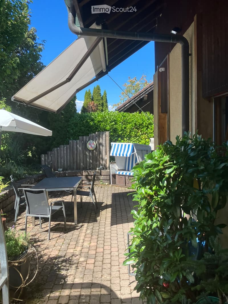 Outdoor patio area with a table, chairs, and an umbrella, surrounded by greenery and a wooden fence.
