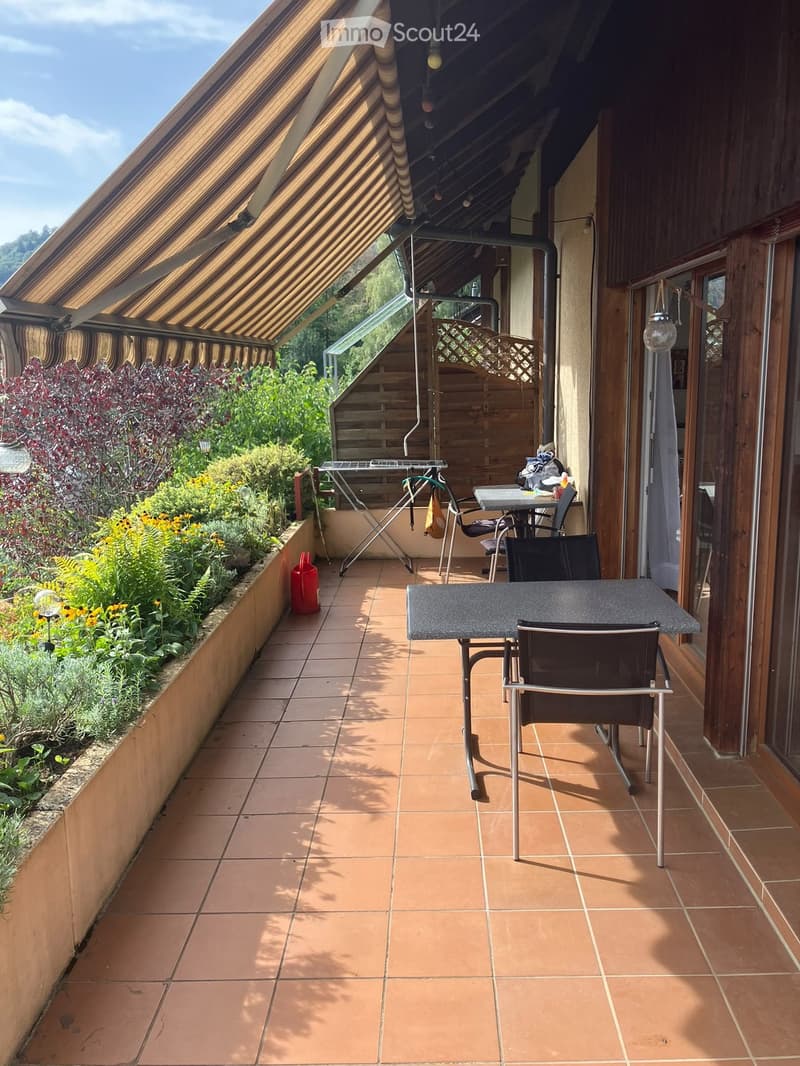 Balcony with tiled floor, wooden railing, and outdoor furniture including a table and chairs. There is a small garden area with plants and flowers on one side.