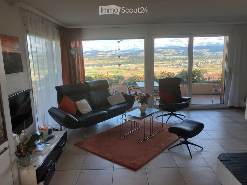 Modern living room with large windows offering a scenic view, featuring a black leather sofa, coffee table, armchair, ottoman, and decorative items.