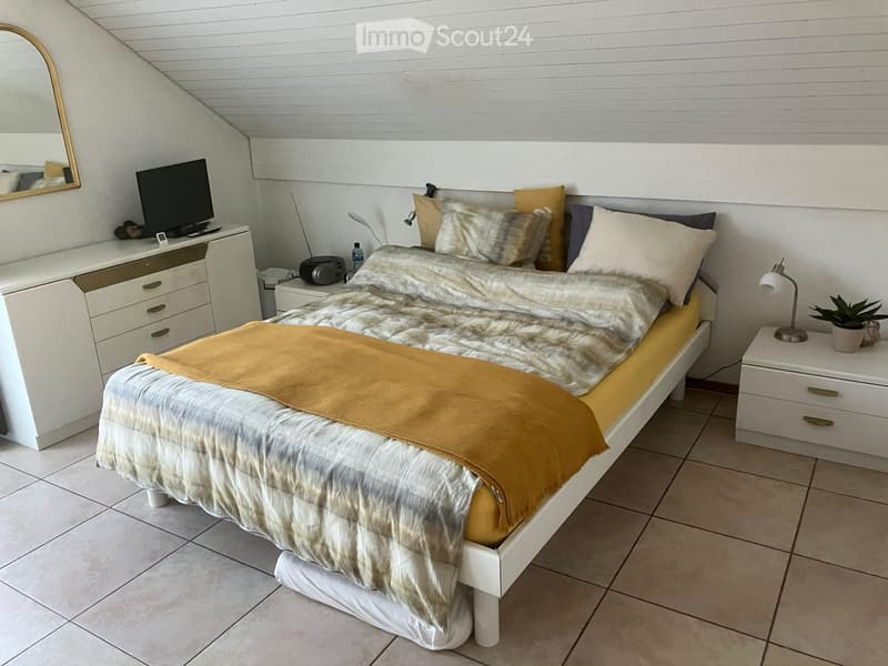 Cozy attic bedroom with a large bed, nightstands, a mirror, and a small potted plant. The room features a slanted ceiling and warm lighting.