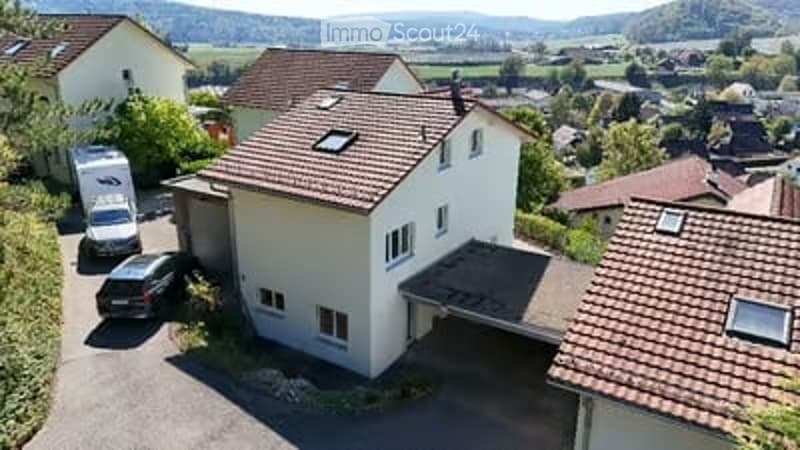 Aerial view of a two-story house with a red-tiled roof, white walls, and a garage. A car is parked in the driveway, and there is a view of surrounding houses and countryside.