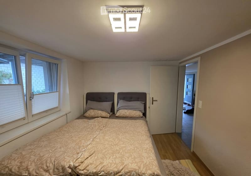 A small, well-lit bedroom with a double bed, headboard, pillows, and a window with blinds. The room features wooden flooring and a door leading to another area.