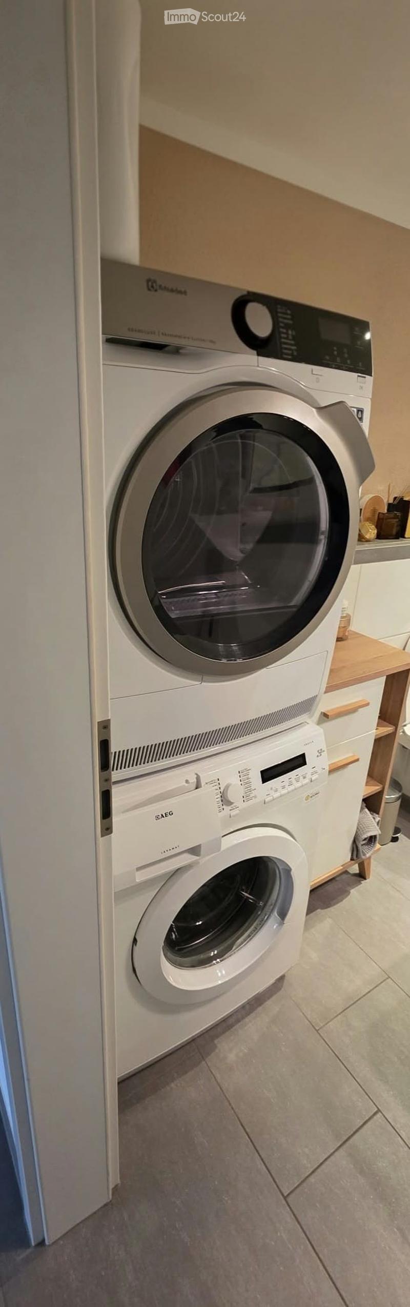 A modern bathroom with a white washing machine and a white dishwasher stacked vertically, with a wooden shelf and small items on the right side.