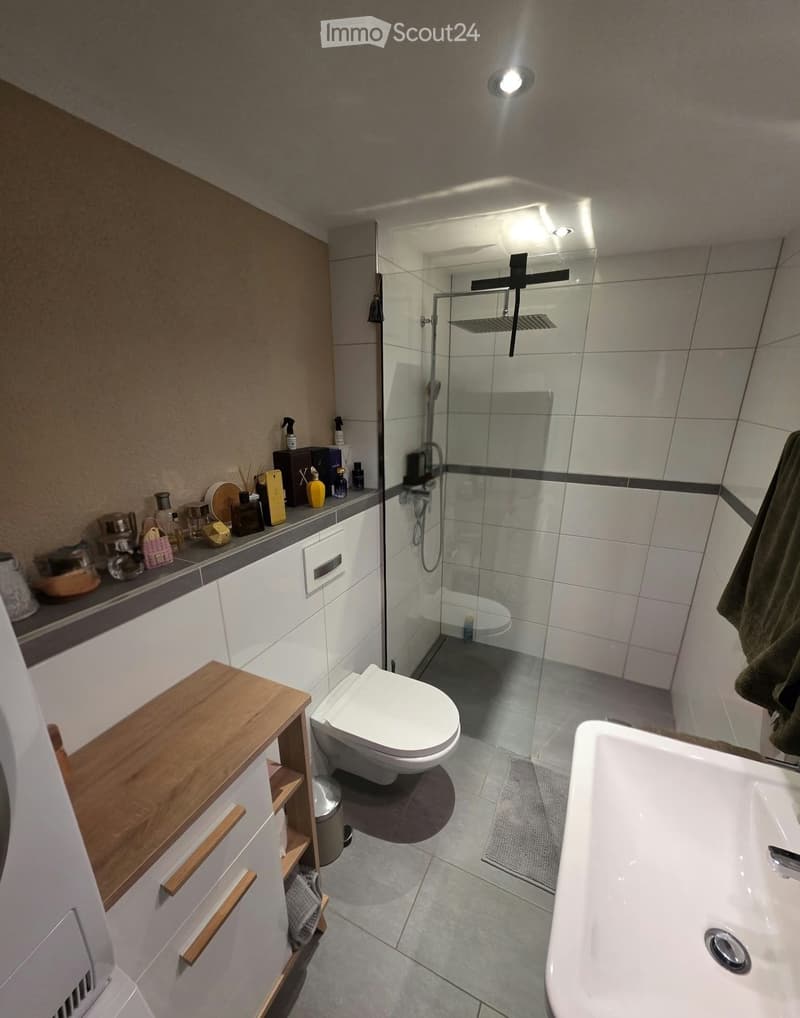 Modern bathroom with a toilet, bathtub, shower, sink, and cabinet. The shower has a glass enclosure, and the sink has a mirror above it. There are various toiletries on the sink counter, and a towel is hanging on the side.