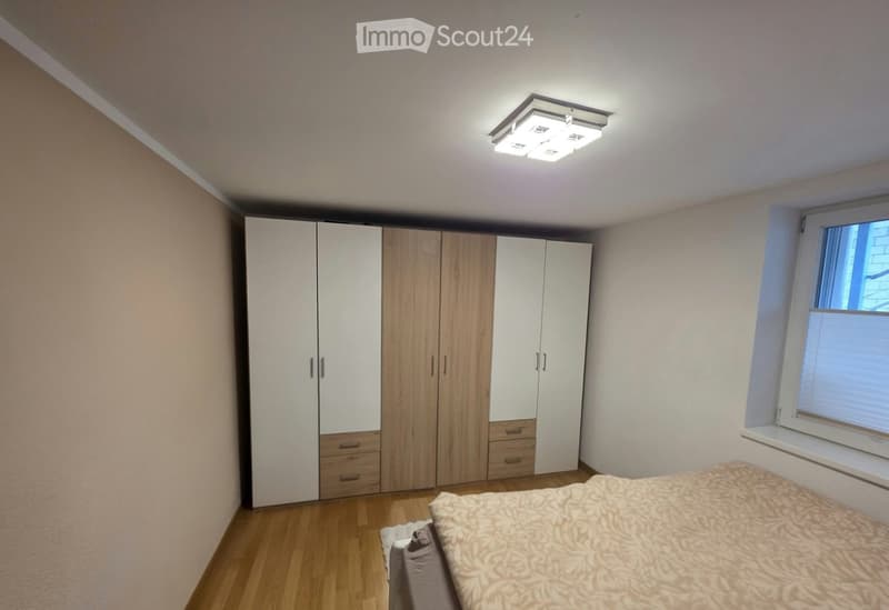 A modern bedroom with a large bed, a tall wardrobe with multiple doors and drawers, and a ceiling light fixture. The room features wooden flooring and a window with blinds.