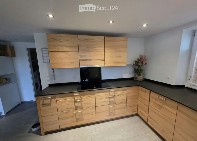 Modern kitchen with light wooden cabinets, black countertops, and stainless steel appliances. Features include a built-in oven, a vase with flowers on the counter, and recessed ceiling lighting.
