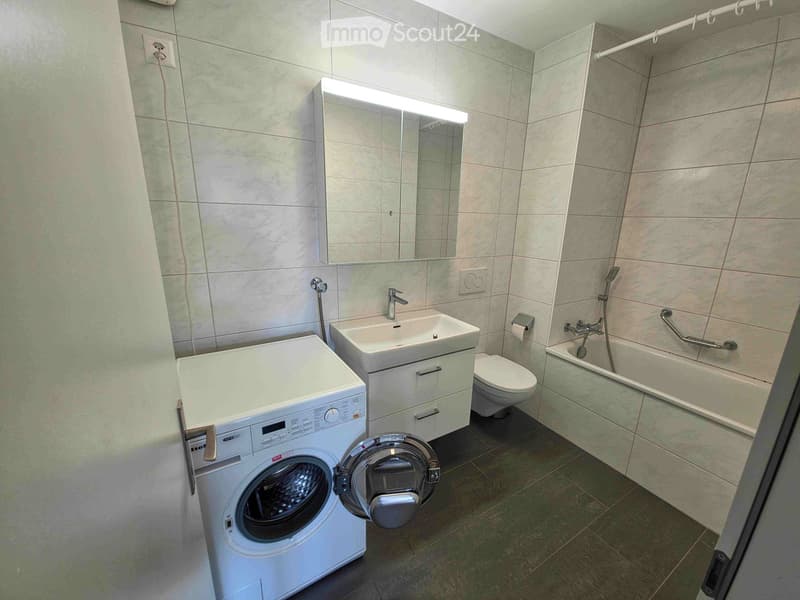 Modern bathroom with a washing machine, sink, toilet, bathtub, and a large mirror with built-in lighting.