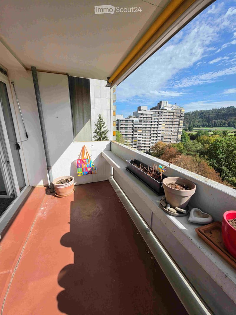 Balcony with terracotta floor, potted plants, and a view of surrounding buildings and greenery.