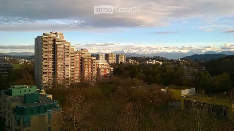 A panoramic view of a cityscape with multiple residential buildings, surrounded by trees and distant mountains under a partly cloudy sky.