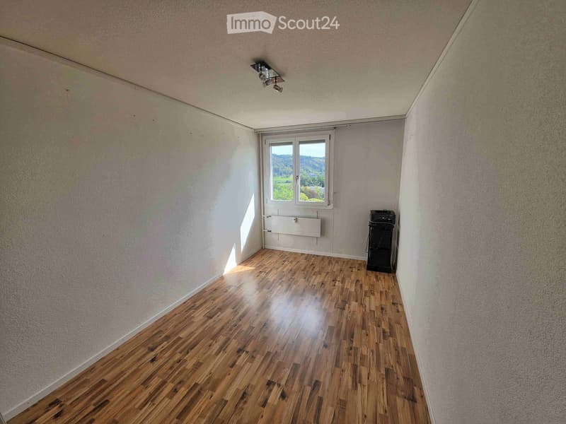Empty room with wooden flooring, white walls, and a window with a view of greenery outside. A small black appliance is placed near the window.