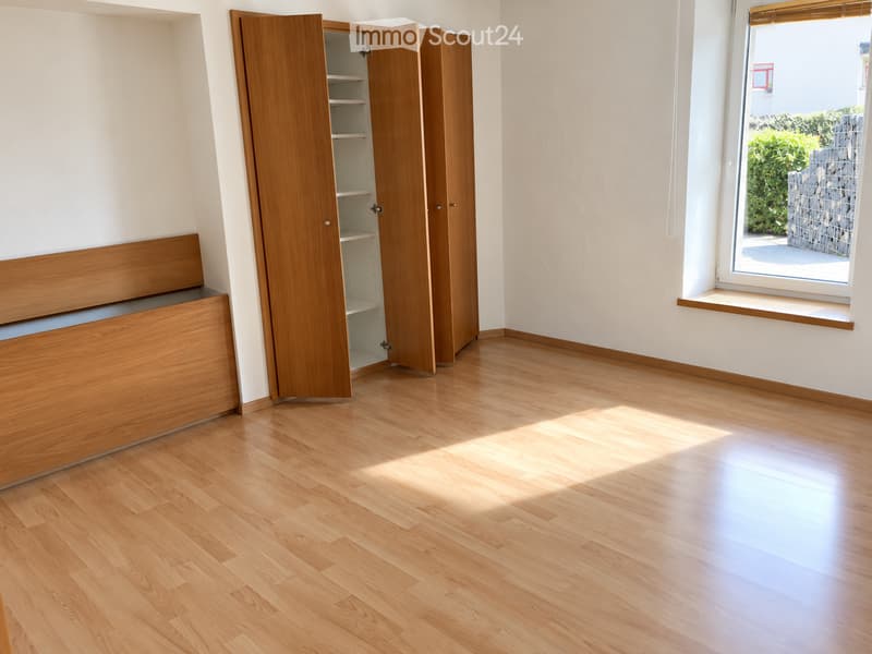 Empty room with light wooden flooring, white walls, built-in wooden bench seating, and a large window with a view of greenery outside.
