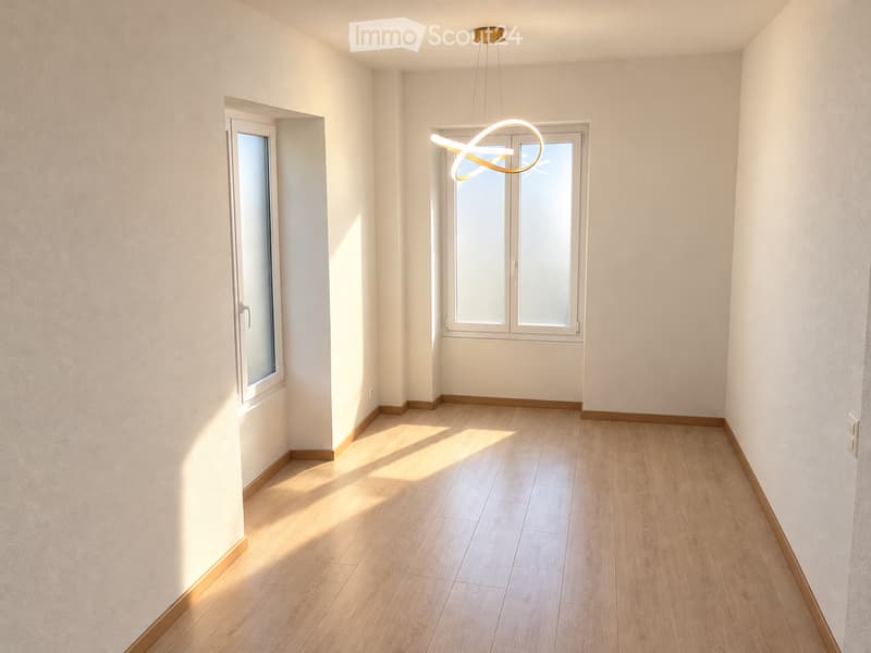 Empty room with light wooden flooring, white walls, and large windows allowing natural light to enter. The room features a modern pendant light fixture hanging from the ceiling.