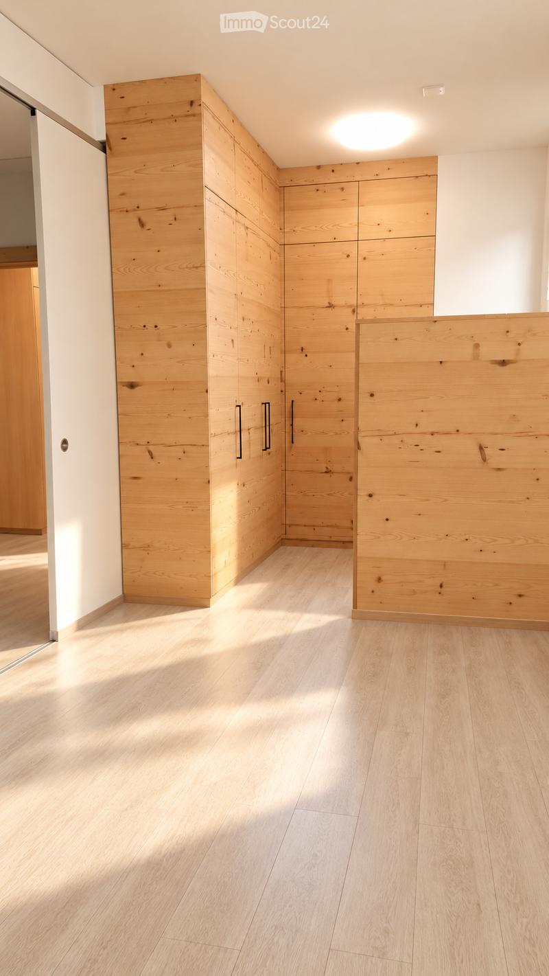 Empty room with light wooden flooring and built-in wooden storage units along one wall. The space is well-lit with natural light coming from an adjacent area.