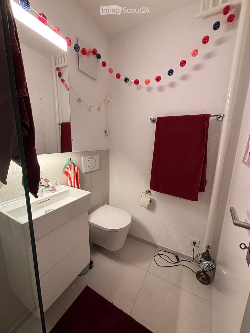 A compact bathroom with a white toilet, sink, and mirror. Red towels are hanging on a rack, and a decorative lamp is placed on the floor. The walls are white, and the floor is tiled.