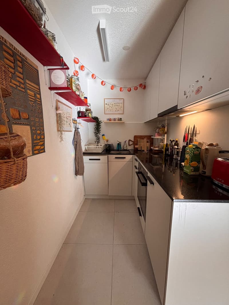 Modern kitchen with white cabinets, black countertop, and various appliances including an oven, toaster, and fridge. Shelves hold jars, a plant, and decorative items.
