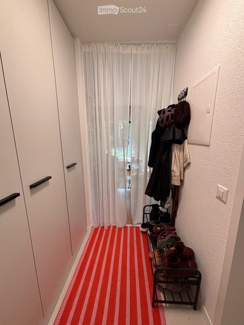 Narrow hallway with white walls, red and white striped carpet, white curtains covering a window at the end, and a coat rack with jackets and shoes on the right side.