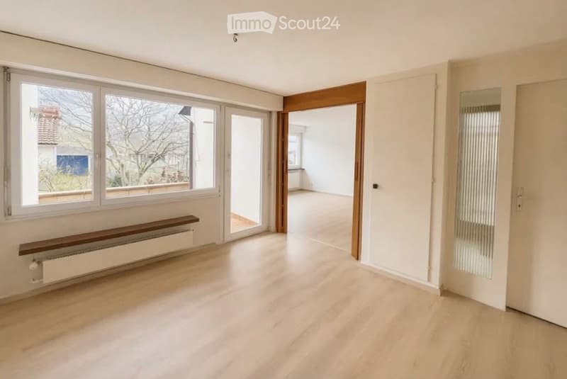 Empty room with light wooden flooring, white walls, large windows, and a wooden bench along one wall.