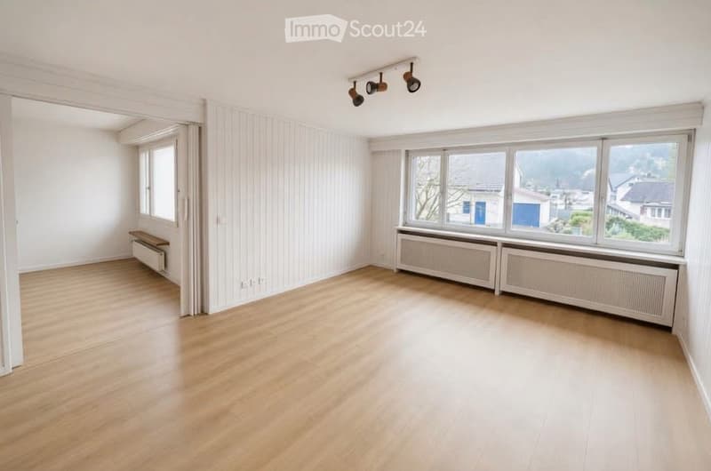 Empty room with wooden flooring, white walls, large windows, radiators, and track lighting.
