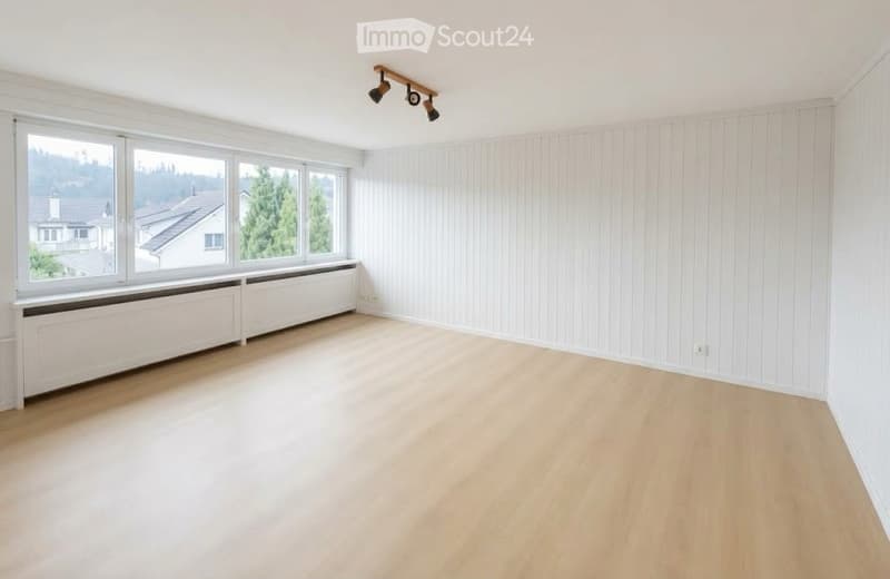Empty room with light wooden flooring, white paneled walls, large windows with white frames, and a simple ceiling light fixture.