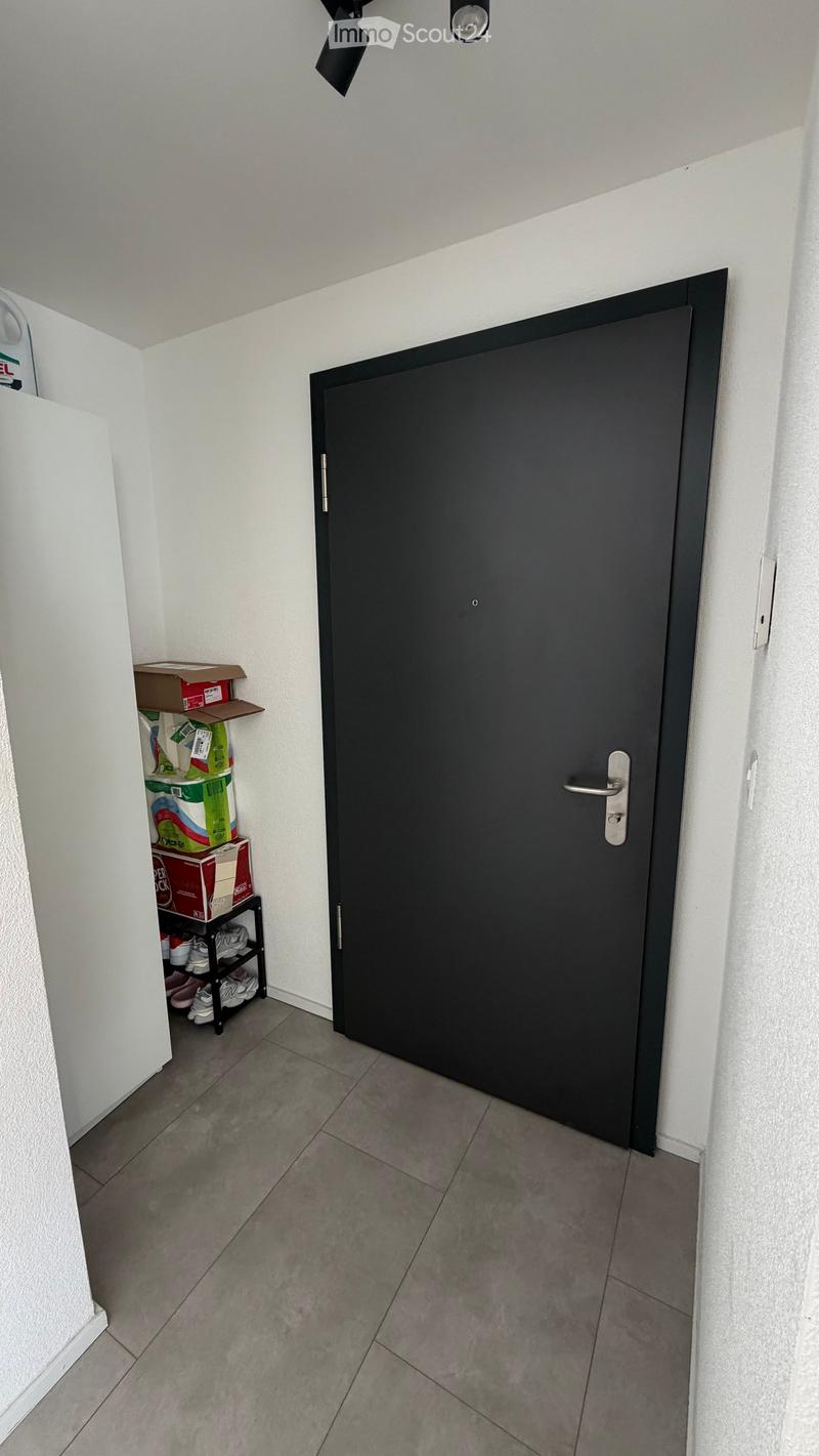 A narrow hallway with a closed black door on the right and a small shelf with boxes and shoes on the left. The floor is tiled, and the walls are white.