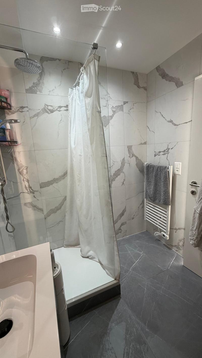 Modern bathroom with marble tiles, a bathtub, a glass-enclosed shower, a towel on a radiator, and a shelf with toiletries.