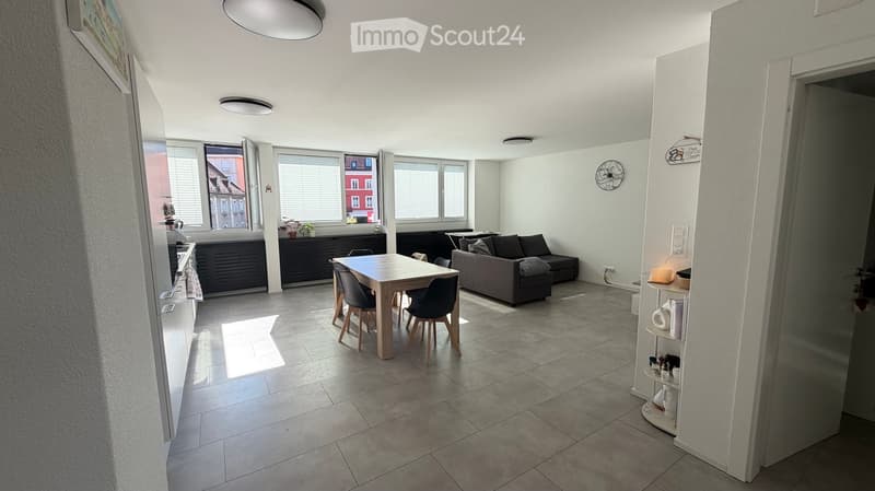 Open-plan living and dining area with a grey sofa, wooden dining table, four chairs, kitchen cabinet, and a small shelf with decorative items.