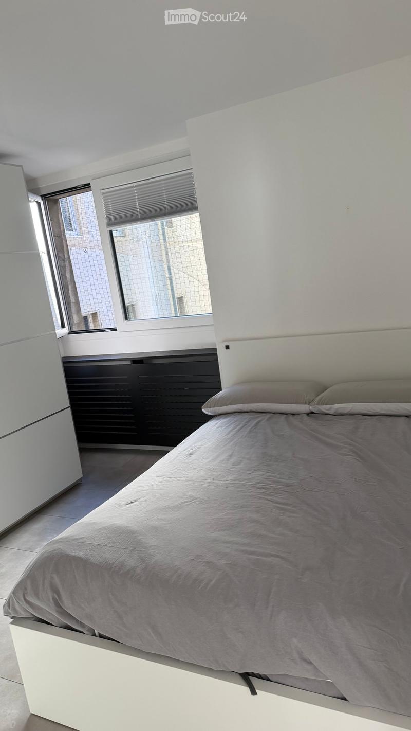 Modern bedroom with a white bed frame, grey bedding, and large windows with blinds.