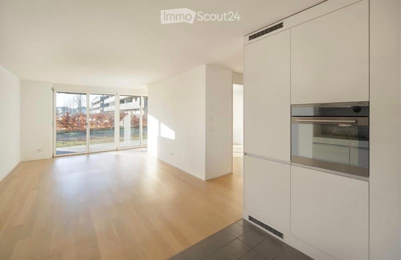Bright, empty room with light wooden flooring, white walls, and large glass sliding doors leading to an outdoor area. Minimalist design with no furniture.
