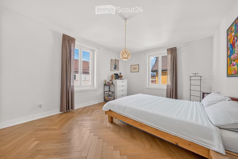Bright bedroom with wooden flooring, large windows with brown curtains, a bed with white linens, a nightstand, and decorative items on shelves.