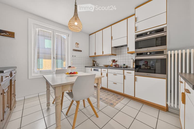 Bright kitchen with white cabinets, a wooden dining table with chairs, a built-in oven, and a window with white curtains.