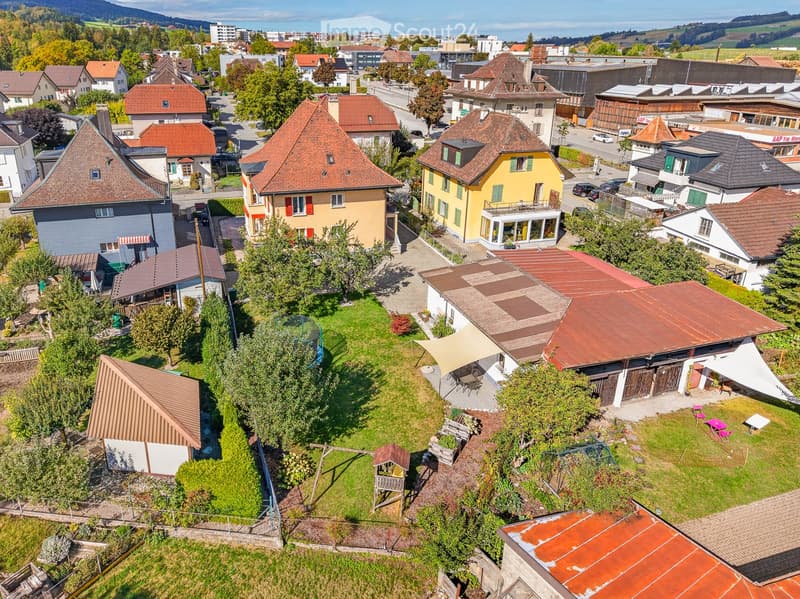 Aerial view of a residential neighborhood with multiple houses, gardens, and outdoor seating areas. The scene includes a mix of architectural styles, green spaces, and a playground.