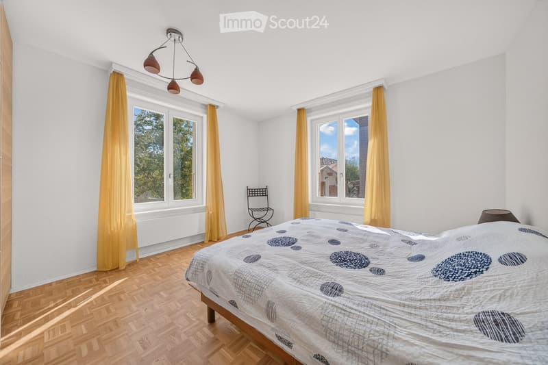 Bright bedroom with wooden flooring, large windows with yellow curtains, a bed with a patterned duvet, a small black chair, and a ceiling light fixture.