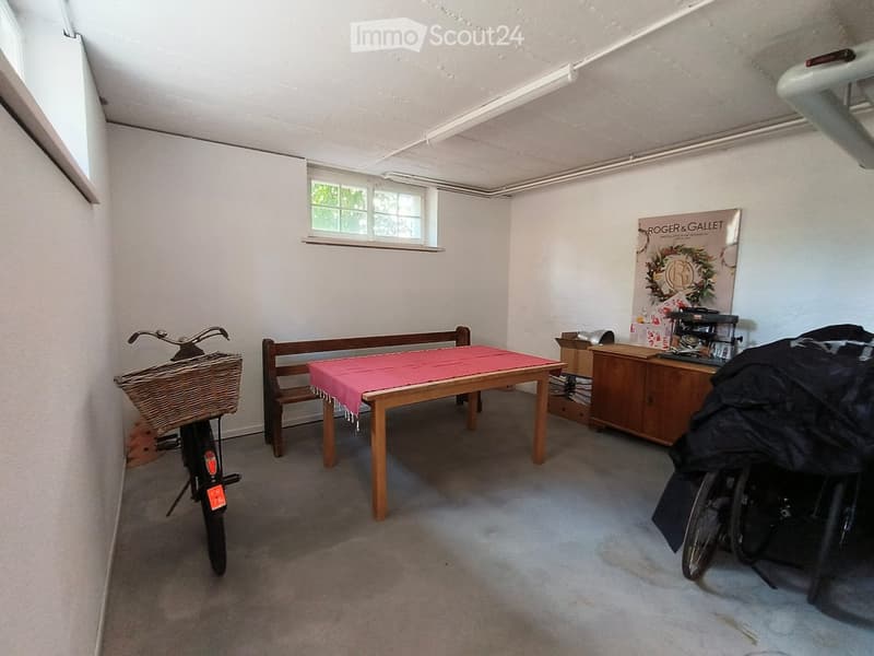 A small, furnished room with a wooden table covered in a red cloth, a bench, a bicycle, a cabinet, and a poster on the wall.