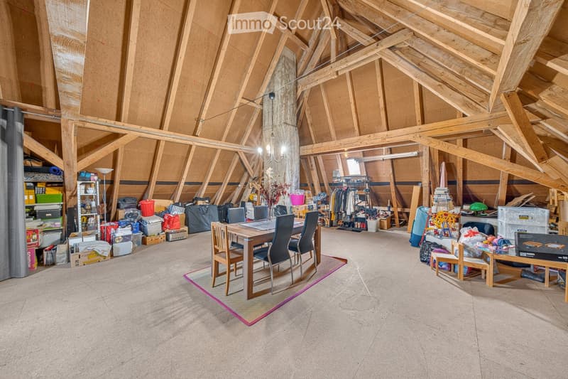 Attic space with exposed wooden beams, concrete floor, and various stored items including a dining table, chairs, shelves, and clothing racks.