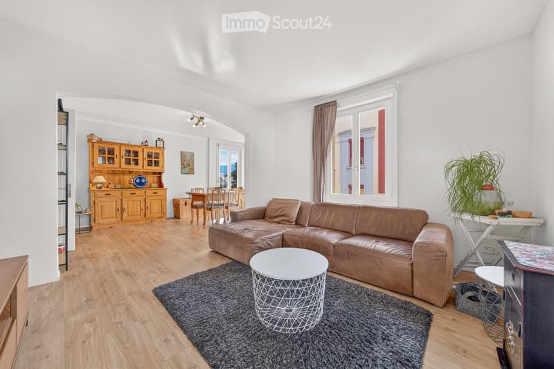 Bright living room with a brown leather sofa, white coffee table, wooden cabinet, dining area with table and chairs, and a potted plant near the window.