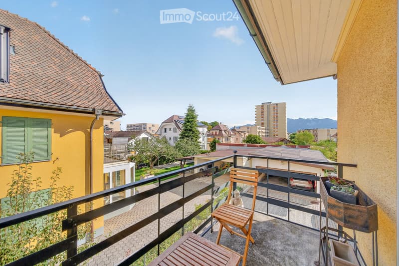 Balcony with wooden chair, table, and potted plant, overlooking a residential area with houses and mountains in the background.