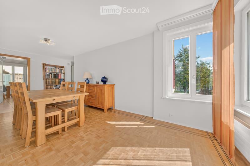 Bright dining room with wooden flooring, large windows, a wooden dining table surrounded by chairs, a wooden cabinet with a lamp and decorative items, and a bookshelf filled with books.