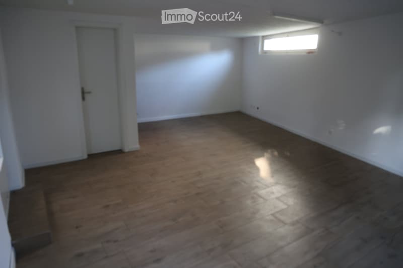 Empty room with wooden flooring, white walls, and a single window letting in natural light. There is a door on the left side.
