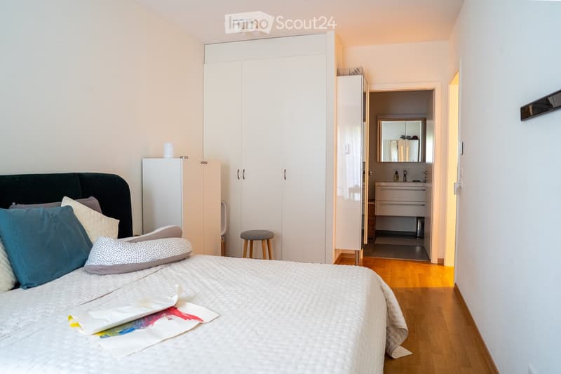 Modern bedroom with a large bed, colorful pillows, a wardrobe, a small stool, and an adjacent bathroom with a sink and mirror.