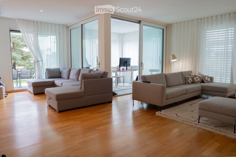 Modern living room with light wooden flooring, large glass sliding doors leading to a balcony, two sectional sofas, ottomans, a small table with chairs, a computer, and a floor lamp.