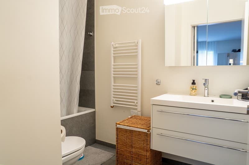 Modern bathroom with a white sink, mirror, toilet, and a wicker basket on the floor. A towel rack and shower curtain are also visible.