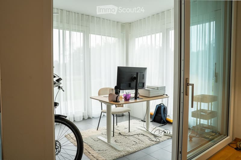 A modern, well-lit room with a white desk, chair, screen, printer, and a bicycle in the corner. The room features large windows with sheer curtains and a glass door leading outside.