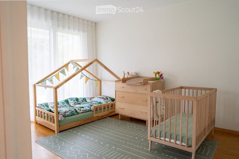 A well-lit children's bedroom with a wooden toy house bed, a crib, a dresser, and a patterned rug.