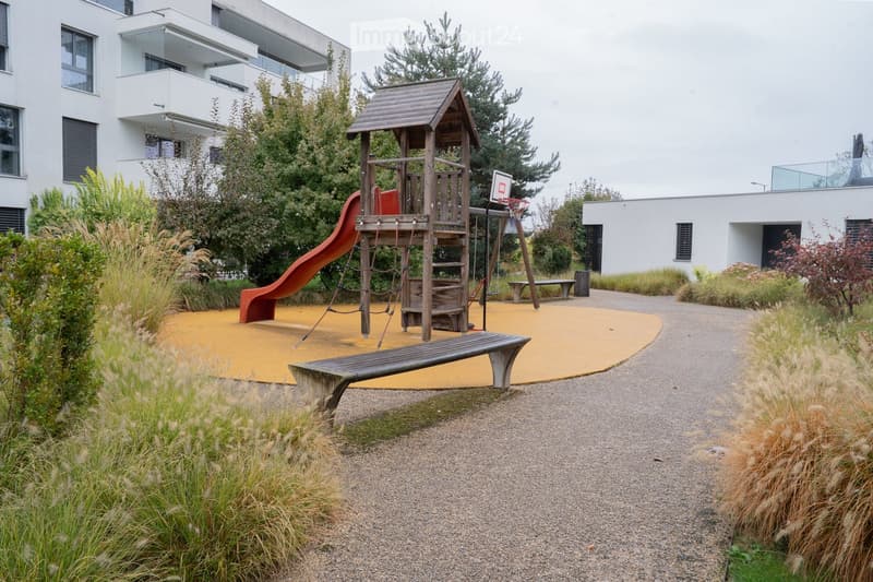 Playground area with a wooden play structure, slide, climbing net, and benches, surrounded by modern apartment buildings and greenery.