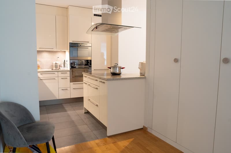 Modern kitchen with white cabinets, stainless steel appliances, and a central island with a granite countertop. A grey chair is placed near the entrance.