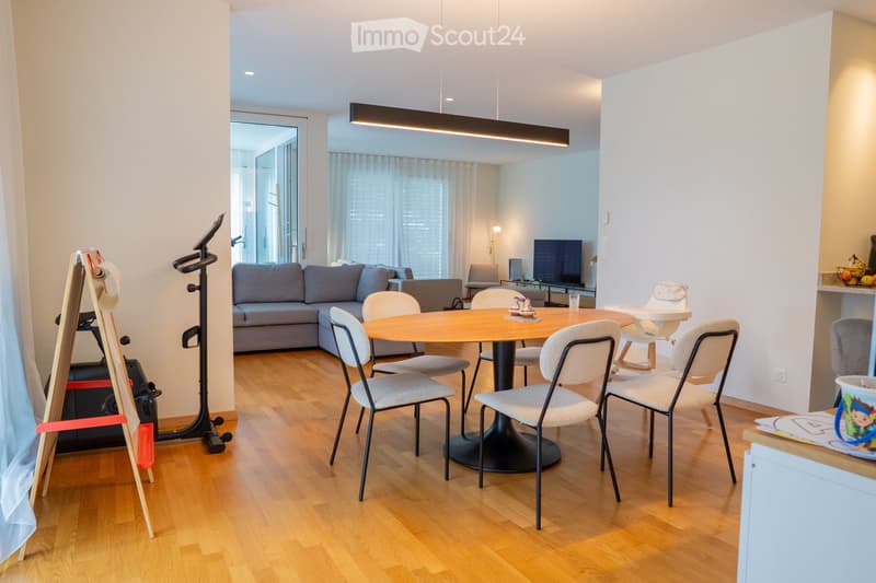 A modern, open-concept living and dining area with a grey sectional sofa, a round wooden dining table with six chairs, a television, and an exercise bike in the corner. The space features light wooden flooring and large windows with white curtains.