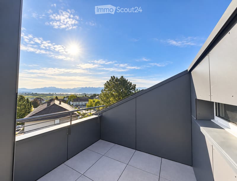 Modern balcony with tiled floor, metal railing, and a scenic view of a countryside landscape with trees and distant mountains under a clear blue sky.