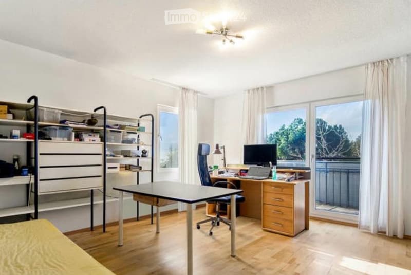 Modern office space with wooden flooring, large windows offering a view, a desk with a laptop, a chair, and a shelf with storage items.