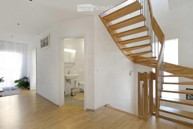A modern, well-lit hallway with a wooden staircase leading upwards, featuring white walls, wooden flooring, and a small adjacent bathroom with a sink and toilet.