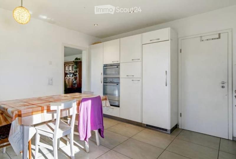 Modern kitchen with white cabinets, stainless steel appliances, a dining table with chairs, and a purple cloth draped over one chair.